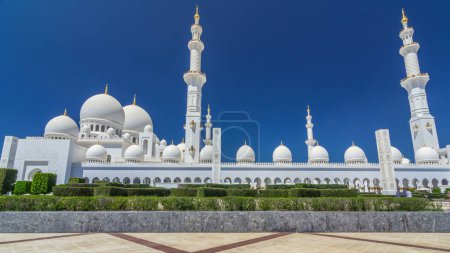 Photo for Sheikh Zayed Grand Mosque timelapse hyperlapse in Abu Dhabi, the capital city of United Arab Emirates. Side view with domes and minarets. Bushes on foreground. Blue sky at sunny day - Royalty Free Image