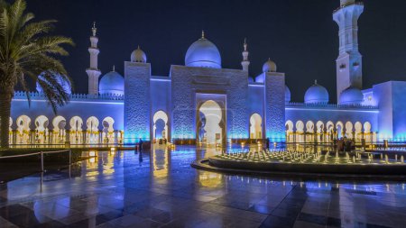 Foto de Sheikh Zayed Gran Mezquita iluminada por la noche hiperlapso timelapse, Abu Dhabi, Emiratos Árabes Unidos. Vista frontal con fuentes. La tercera mezquita más grande del mundo - Imagen libre de derechos