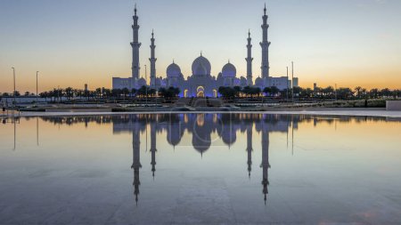 Foto de Sheikh Zayed Gran Mezquita en Abu Dhabi día a noche timelapse transición después de la puesta del sol, Emiratos Árabes Unidos. Vista nocturna desde Wahat Al Karama con reflexiones sobre el agua - Imagen libre de derechos