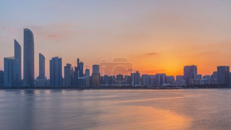 Foto de Horizonte de la ciudad de Abu Dhabi en la hora del amanecer con el tiempo de reflexión del agua desde el rompeolas cerca de la aldea cultural. Pocas nubes en el cielo matutino - Imagen libre de derechos