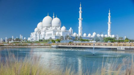 Téléchargez les photos : Timelapse de la Grande Mosquée Cheikh Zayed à Abu Dhabi, la capitale des Émirats arabes unis. Face arrière avec lac et fontaine. Ciel bleu au soleil - en image libre de droit