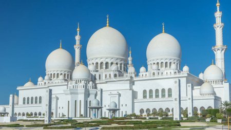 Photo for Sheikh Zayed Grand Mosque timelapse in Abu Dhabi, the capital city of United Arab Emirates. Back side with trees and traffic on the road. Blue sky at sunny day - Royalty Free Image