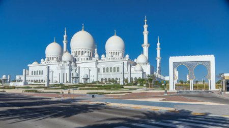 Photo for Sheikh Zayed Grand Mosque timelapse in Abu Dhabi, the capital city of United Arab Emirates. Back side with trees and traffic on the road. Blue sky at sunny day - Royalty Free Image