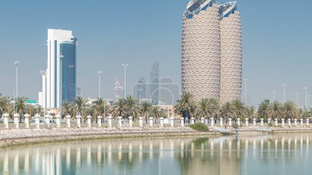 Foto de Vista del horizonte de los rascacielos con las torres Al Bahr en Abu Dhabi timelapse. Reflexiones sobre el agua y las palmas en el paseo marítimo. Emiratos Árabes Unidos - Imagen libre de derechos