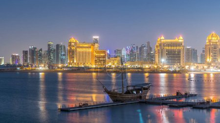 Téléchargez les photos : Vue de Katara Beach du jour à la nuit après le coucher du soleil à Doha, Qatar, vers la baie de l'Ouest et le centre-ville. Des gratte-ciel éclairés. Vieux navire au premier plan - en image libre de droit