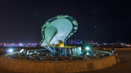 Téléchargez les photos : Corniche avec fontaine ornée d'une huître avec une perle gigantesque à l'intérieur illuminée par une hyperlapse nocturne intemporelle avec l'horizon de Doha et les bateaux derrière. Des gens qui marchent - en image libre de droit
