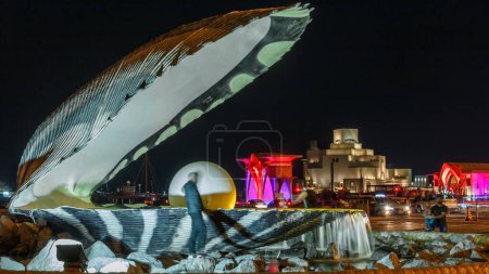 Téléchargez les photos : Corniche avec fontaine ornée d'une huître avec une perle gigantesque à l'intérieur illuminée par le timelapse nocturne avec l'horizon de Doha et le musée derrière. Des gens qui marchent - en image libre de droit