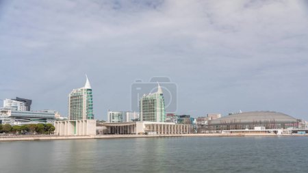 Moderne hohe Wohn- und Bürogebäude, die sich im Wasser unter blauem wolkenverhangenem Himmel spiegeln, überschlagen sich im Zeitraffer in Lissabon, Portugal. Park Nations Luxusviertel mit Konzerthalle und Flaggen