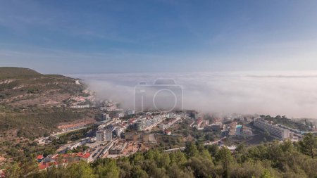 Téléchargez les photos : Panorama montrant la vue aérienne de la ville de Sesimbra et le port couvert par le brouillard timelapse, Portugal. Paysage supérieur au-dessus des nuages du point de vue du château. Resort dans le quartier de Setubal - en image libre de droit
