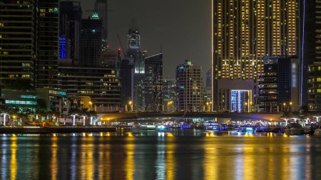 Foto de Paseo marítimo y puente con el tráfico sobre el canal en Dubai Marina timelapse por la noche, Emiratos Árabes Unidos. Vista desde el puente con palmeras, barcos y torres. Distrito con canal artificial y rascacielos iluminados - Imagen libre de derechos