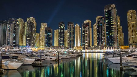 Téléchargez les photos : Vue de nuit depuis la promenade sur Dubai Marina tours éclairées reflétées dans l'eau avec des yachts flottants et des bateaux timelapse hyperlapse, Émirats arabes unis. Quartier de Dubaï et un canal artificiel - en image libre de droit