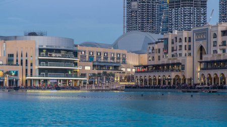 Téléchargez les photos : La fontaine dansante de Dubaï et le timelapse du lac du jour au soir se produisent au rythme de la musique sélectionnée au crépuscule à Dubaï, Émirats arabes unis. La fontaine est dominée par le centre commercial et les Hôtels - en image libre de droit