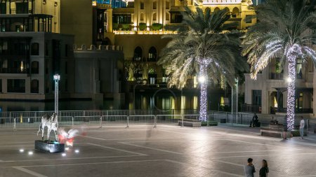 Place avec monument équestre près de Souk dans le quartier du centre-ville timelapse à Dubaï, EAU Palmiers et bâtiments illuminés et reflétés dans l'eau du lac