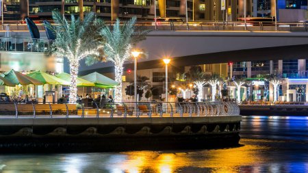 Foto de Paseo marítimo con cafeterías y restaurantes en Dubai Marina timelapse por la noche, Emiratos Árabes Unidos. Vista desde el paseo marítimo con palmeras, barcos y torres. Distrito con canal artificial - Imagen libre de derechos
