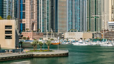 Foto de Dubai Marina paseo marítimo, palmeras, modernas torres con yates flotantes y barcos de puente timelapse, Emiratos Árabes Unidos. Canal artificial y rascacielos alrededor - Imagen libre de derechos