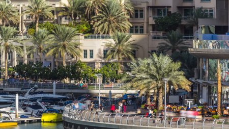 Téléchargez les photos : Vue rapprochée sur la promenade de Dubai Marina, les palmiers, les tours modernes avec des yachts flottants et des bateaux depuis le pont Timelapse, Émirats arabes unis. Personnes marchant près des cafés - en image libre de droit