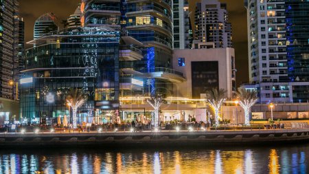 Téléchargez les photos : Canal de la marina de Dubaï avec des bateaux et des ponts au-dessus, des palmiers et des tours modernes reflétés dans l'eau du front de mer à Dubaï timelapse de nuit, Émirats arabes unis. Des gratte-ciel éclairés autour - en image libre de droit