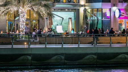 Foto de Paseo marítimo con cafeterías y restaurantes en Dubai Marina timelapse por la noche, Emiratos Árabes Unidos. Vista desde el paseo marítimo con palmeras, barcos y torres. Distrito con canal artificial - Imagen libre de derechos