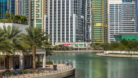 Foto de Timelapse vista sobre rascacielos en el paseo marítimo con palmeras. Edificios residenciales en Jumeirah Lake Towers reflejados en el agua en Dubai, Emiratos Árabes Unidos. Vista con agua verde desde el paseo marítimo - Imagen libre de derechos