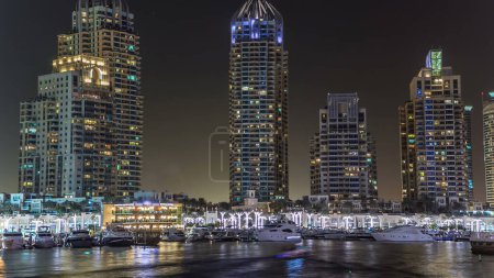 Foto de Paseo marítimo y canal en Dubai Marina timelapse por la noche, Emiratos Árabes Unidos. Vista aérea desde puente con palmeras, barcos flotantes y torres iluminadas. Puerto con yates - Imagen libre de derechos