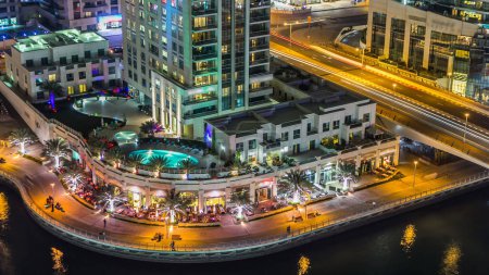 Foto de Vista aérea en la noche del paseo marítimo de Dubai Marina con restaurantes y canal con yates flotantes y barcos en Dubai, Emiratos Árabes Unidos. Torres modernas iluminadas con luces y tráfico en el puente - Imagen libre de derechos