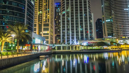 Foto de Timelapse vista nocturna en rascacielos iluminados en el paseo marítimo con palmeras. Edificios residenciales en Jumeirah Lake Towers en Dubai, Emiratos Árabes Unidos. Vista desde el paseo marítimo - Imagen libre de derechos
