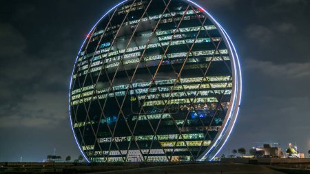 Photo for The Aldar headquarters building night timelapse with glowing windows is the first circular building of its kind in the Middle East in Abu Dhabi, UAE. Cloudy sky - Royalty Free Image