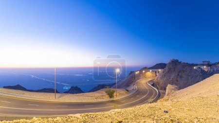 Tag-Nacht-Übergangszeit mit Felsen und kurviger Straße am Jebel Hafeet. Der Berg liegt hauptsächlich in der Umgebung von Al Ain und bietet einen beeindruckenden Blick über die Stadt. Luftaufnahme von oben