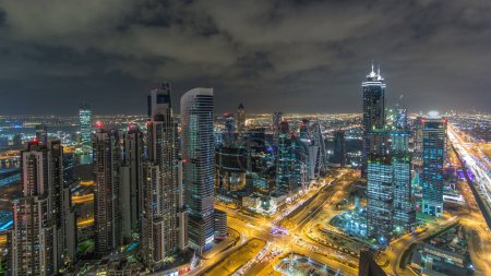 Foto de Dubai torres de la bahía de negocios iluminadas por la noche timelapse. Tráfico en carretera con intersección. Vista aérea en la azotea de algunos rascacielos y nuevas torres en construcción. - Imagen libre de derechos