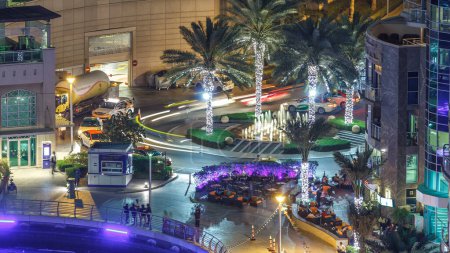 Foto de Dubai Marina fountain aerial night timelapse, restaurants and marina promenade, Dubai, Emiratos Árabes Unidos. Palmeras iluminadas y el tráfico en una carretera circular con taxi - Imagen libre de derechos