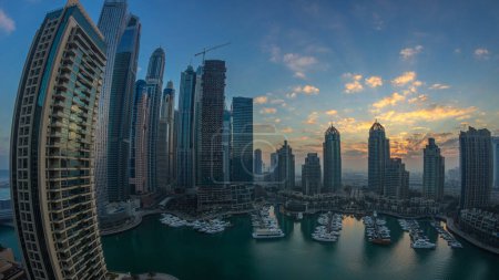 Téléchargez les photos : Panorama des gratte-ciel modernes qui brillent dans les lumières du lever du soleil timelapse à Dubai Marina avec des yachts à Dubaï, Émirats arabes unis. Rayons de lumières derrière les bâtiments. Nuages orange sur le ciel - en image libre de droit
