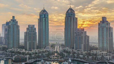 Foto de Vista de rascacielos modernos que brillan al amanecer luces timelapse en Dubai Marina waterfront con yates en Dubai, Emiratos Árabes Unidos. Rayos de luces detrás de edificios. Nubes naranjas en el cielo - Imagen libre de derechos