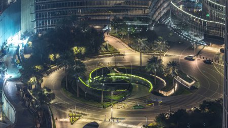 Dubai traffic on circle at night timelapse with beautiful fountain in the center with lights close to skyscraper. Aerial top view with palms and trees