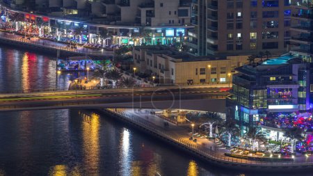 Foto de Canal de agua y paseo marítimo en Dubai Marina horizonte por la noche timelapse. Torres residenciales con iluminación e iluminación. Yates y barcos flotantes con tráfico en un puente - Imagen libre de derechos