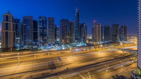 Die Skyline der Jumeirah-Seen aus der Luft mit ihren beleuchteten Wolkenkratzern übergeht Nacht auf Tag mit dem Verkehr auf der Scheich-Zayed-Straße und der U-Bahn-Linie. Blick auf die Dächer von Dubais Yachthafen vor Sonnenaufgang
