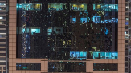 Windows of the multi-storey building of glass and steel lighting inside and moving people within timelapse. Aerial view of modern office skyscrapers in Dubai.