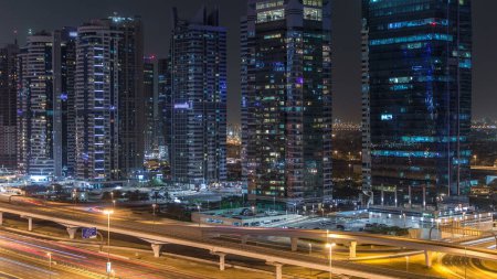 Foto de Vista aérea de las torres de los lagos Jumeirah con rascacielos iluminados timelapse noche con el tráfico en la carretera zayed jeque y la línea de metro. Vista desde el puerto deportivo de Dubái - Imagen libre de derechos