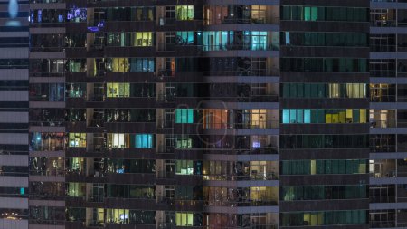 Téléchargez les photos : Fenêtres du bâtiment de plusieurs étages d'éclairage en verre et en acier à l'intérieur et les personnes en mouvement dans le temps. Vue aérienne des gratte-ciel résidentiels modernes à Dubaï - en image libre de droit