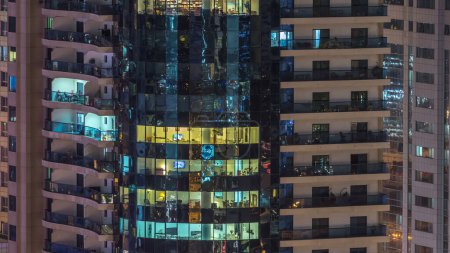Foto de Ventanas del edificio de varios pisos de iluminación de vidrio y acero en el interior y personas en movimiento dentro de timelapse. Vista aérea de los modernos rascacielos residenciales y de oficinas en Dubai marina - Imagen libre de derechos