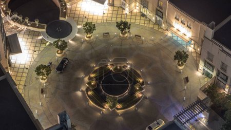 Dubai traffic on circle at night timelapse with fountain in the center with lights between skyscrapers. Aerial top view with trees in yard