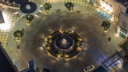 Dubai traffic on circle at night timelapse with fountain in the center with lights between skyscrapers. Aerial top view with trees