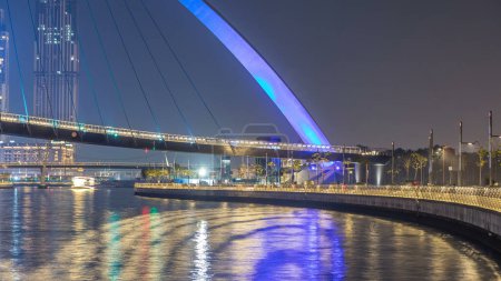 Téléchargez les photos : Pont piétonnier futuriste au-dessus du canal d'eau de Dubaï Illuminé la nuit timelapse, EAU. Paysage urbain avec front de mer en arrière-plan. Vue de la promenade - en image libre de droit