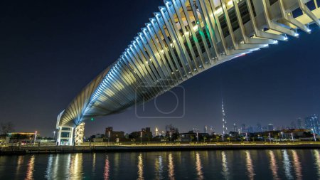 Téléchargez les photos : Pont piétonnier tordu futuriste au-dessus du canal d'eau de Dubaï Illuminé la nuit hyperlapse timelapse, EAU. Paysage urbain en arrière-plan. Vue de la promenade - en image libre de droit