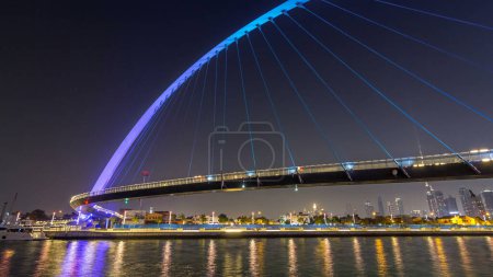 Photo for Futuristic Pedestrian Bridge over the Dubai Water Canal Illuminated at Night timelapse hyperlapse, UAE. Cityscape on background. View from promenade - Royalty Free Image