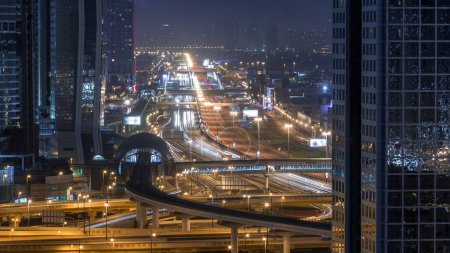 Foto de Dubai skyline céntrico noche a día timelapse transición y Sheikh Zayed tráfico de intersección de carreteras, Emiratos Árabes Unidos. Vista aérea desde la azotea del rascacielos con torres modernas iluminadas y línea de metro - Imagen libre de derechos