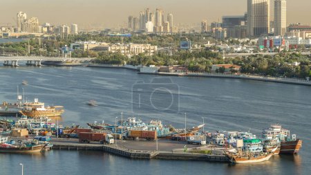 Téléchargez les photos : Dubaï Creek paysage timelapse avec des bateaux et des navires dans le port, l'eau calme et des bâtiments modernes en arrière-plan pendant le coucher du soleil. Vue aérienne du dessus d'en haut - en image libre de droit