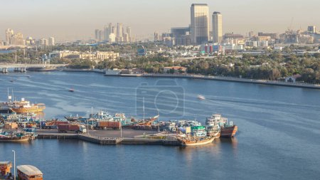 Téléchargez les photos : Dubaï Creek paysage timelapse avec des bateaux et des navires dans le port, l'eau calme et des bâtiments modernes en arrière-plan pendant le coucher du soleil. Vue aérienne du dessus de la jetée d'en haut - en image libre de droit