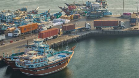 Téléchargez les photos : Chargement d'un navire à Port Said timelapse à Dubaï, Deira creek, EAU. Camions avec conteneurs. Vue aérienne du dessus avec de nombreux vieux bateaux au coucher du soleil avec une lumière chaude - en image libre de droit