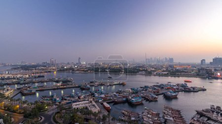 Dubai creek landscape panorama day to night transition timelapse with boats and yachts and modern buildings with traffic on the road. Aerial top view from above