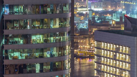 Photo for Windows of the multi-storey building of glass and steel lighting inside and moving people within timelapse. Aerial view of modern office skyscrapers in Dubai deira. Water of creek on background - Royalty Free Image